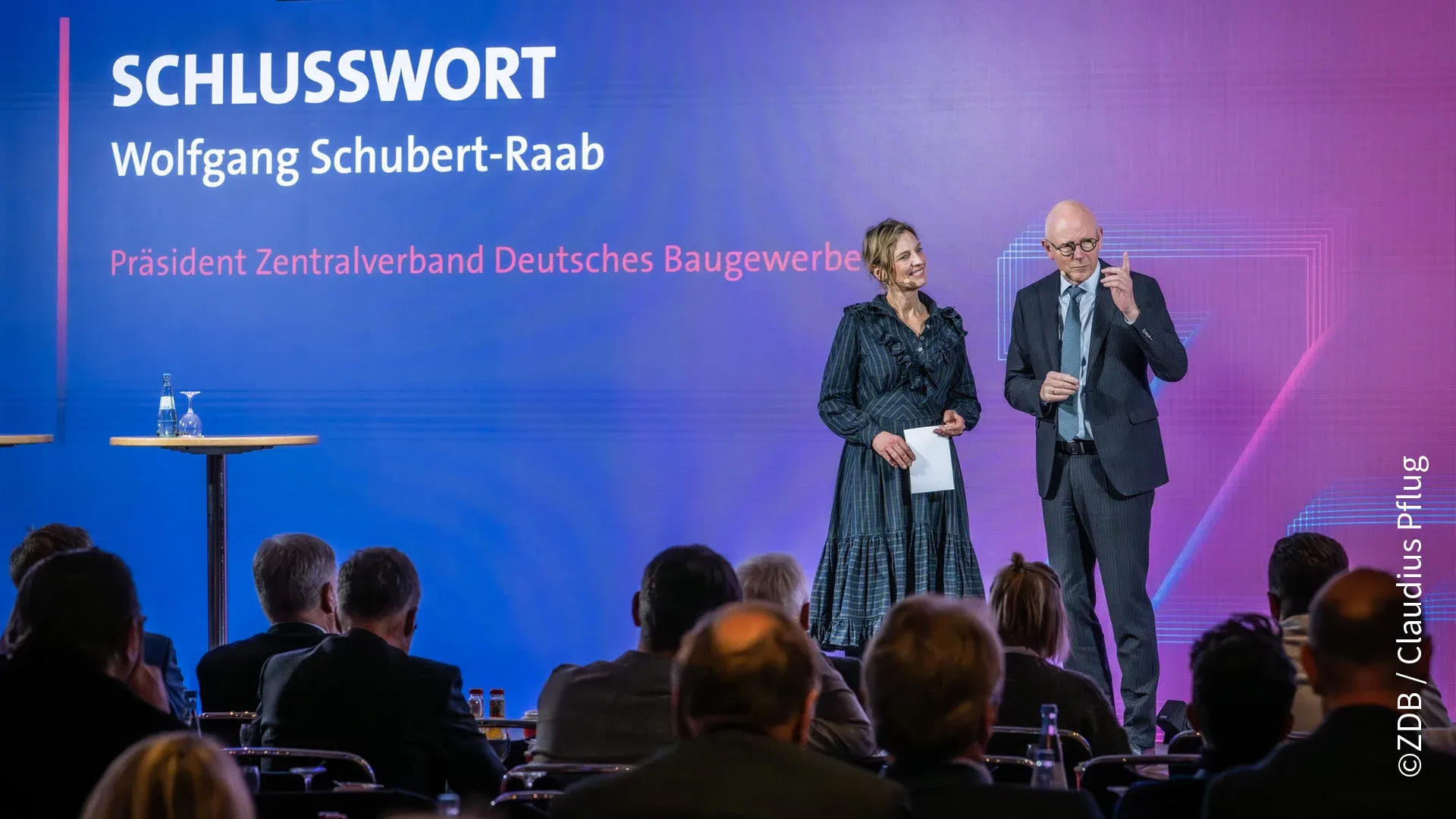 Zwei Personen sprechen auf der Bühne vor der gestalteten Rückwand 