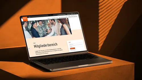 Die Login-Seite zum Mitgliederbereich der RAL Gütezeichen auf einem Laptop vor orangefarbenem Hintergrund, mit einem Foto lachender Personen im Businessumfeld und einem Anmeldeformular.